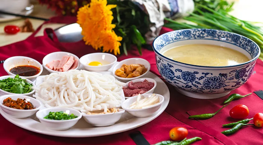 «Лапша, переходящая мост» (Crossing-the-bridge noodles) — это юньнаньское блюдо, где в миску с кипящим бульоном, запечатанным слоем куриного жира, прямо за столом добавляют сырые тонкие ломтики мяса, рыбы, перепелиные яйца и рисовую лапшу